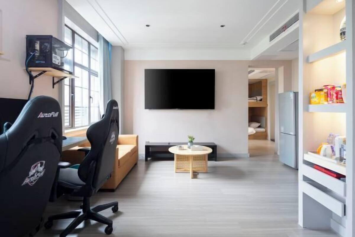 Een moderne woonkamer met een gaming stoel, een flatscreen televisie, een bank, een klein rond tafeltje en een keuken met een koelkast.