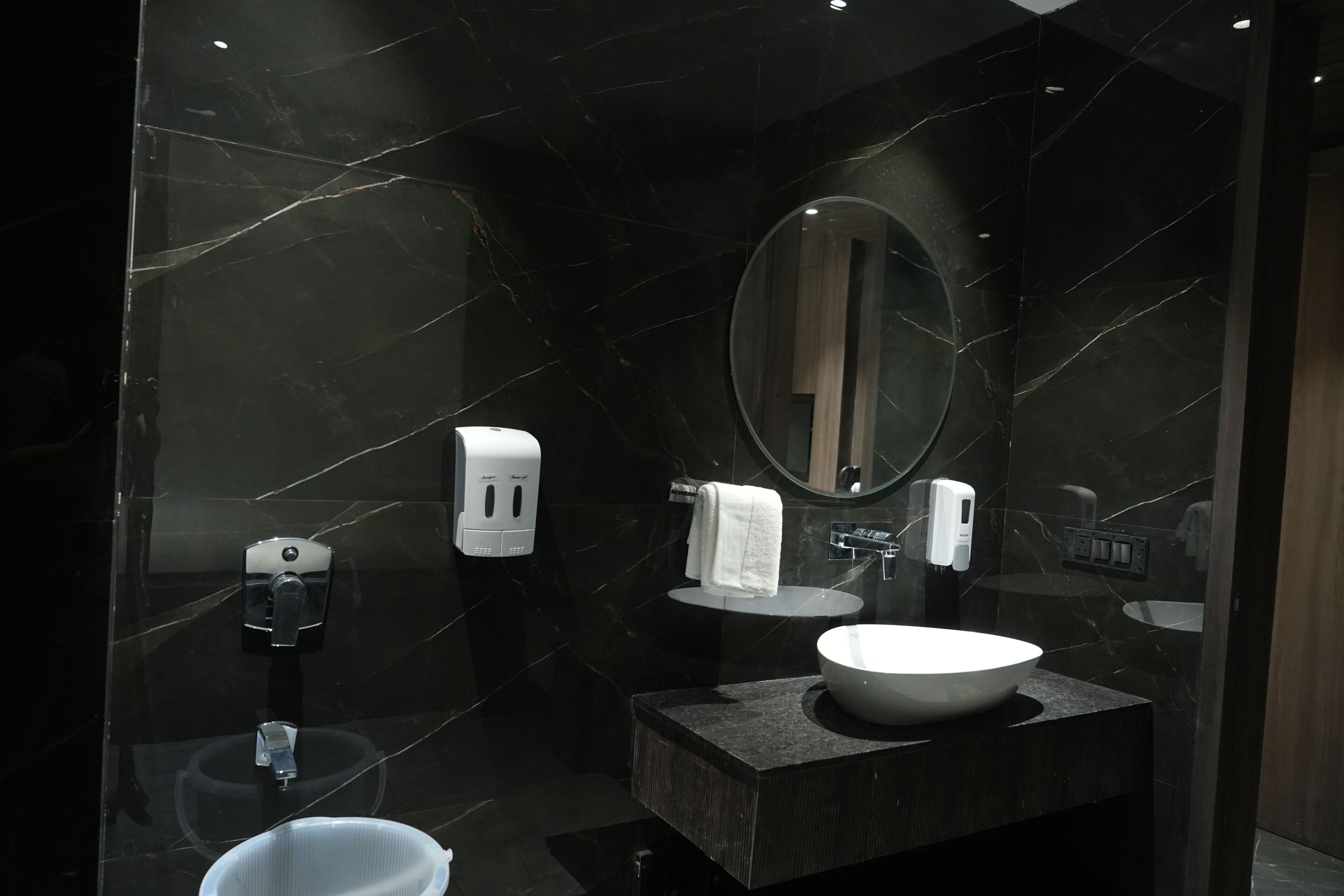 Un baño moderno con pared de mármol, un espejo redondo, un lavamanos, dispensador de toallas y un inodoro.