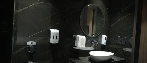 Un baño moderno con pared de mármol, un espejo redondo, un lavamanos, dispensador de toallas y un inodoro.