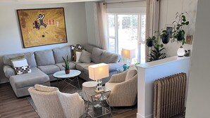 A spacious living room with a grey sofa, a round coffee table, and a beige armchair. There is a painting on the wall and a radiator near the window.