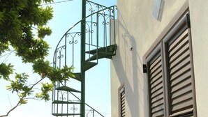 Een spiraaltrap met een metalen leuning die naar een balkon leidt, een bruine gesloten deur en uitzicht op de zee.