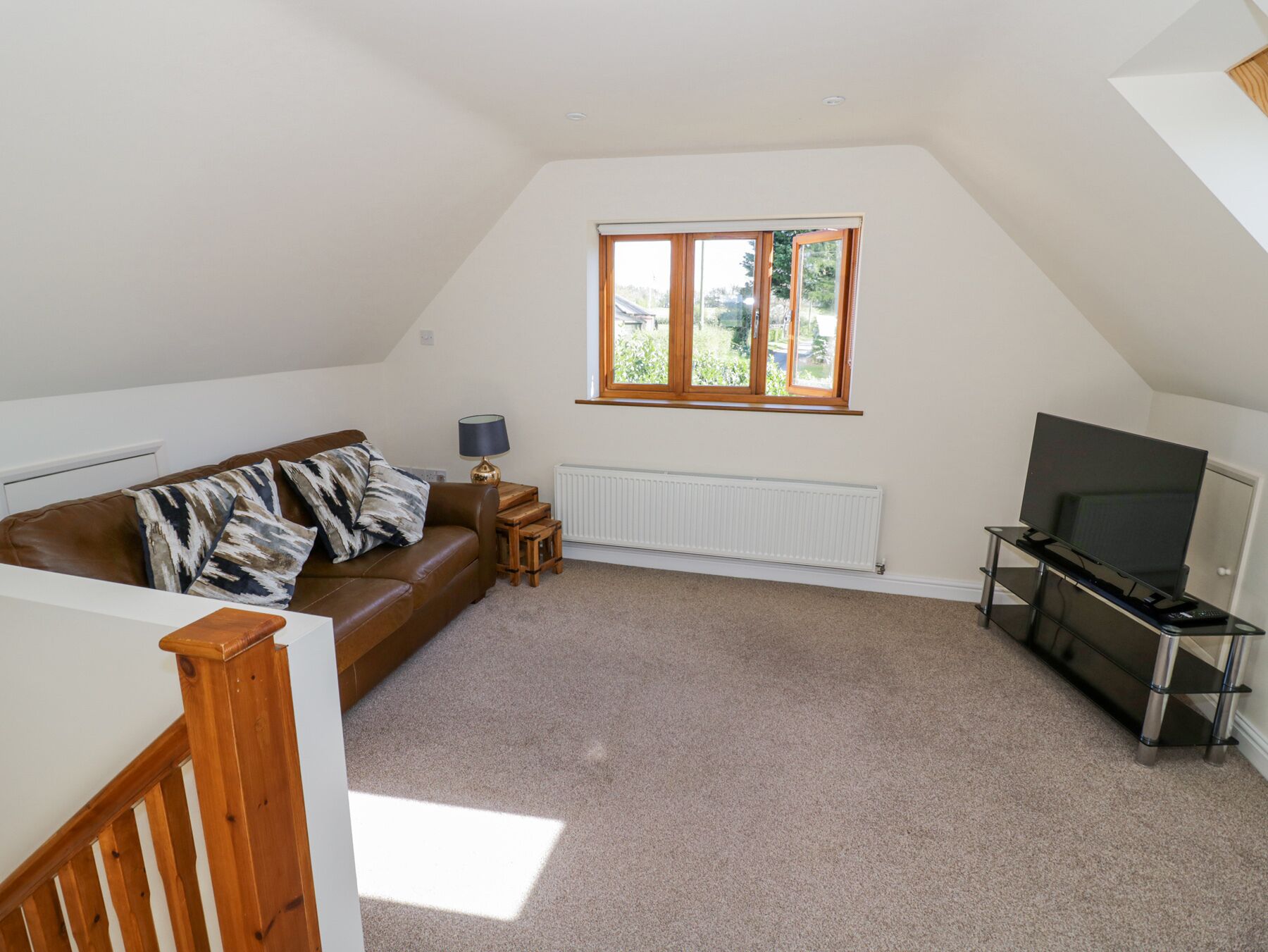 A spacious living room with a brown leather sofa, a TV on a glass stand, a window with a view of greenery, and a radiator.