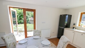 A dining area with a round table set for four, surrounded by floral-patterned chairs. A black refrigerator is visible in the background, and there is a window with a view of greenery outside. - Little Barn (Stratford-upon-Avon)