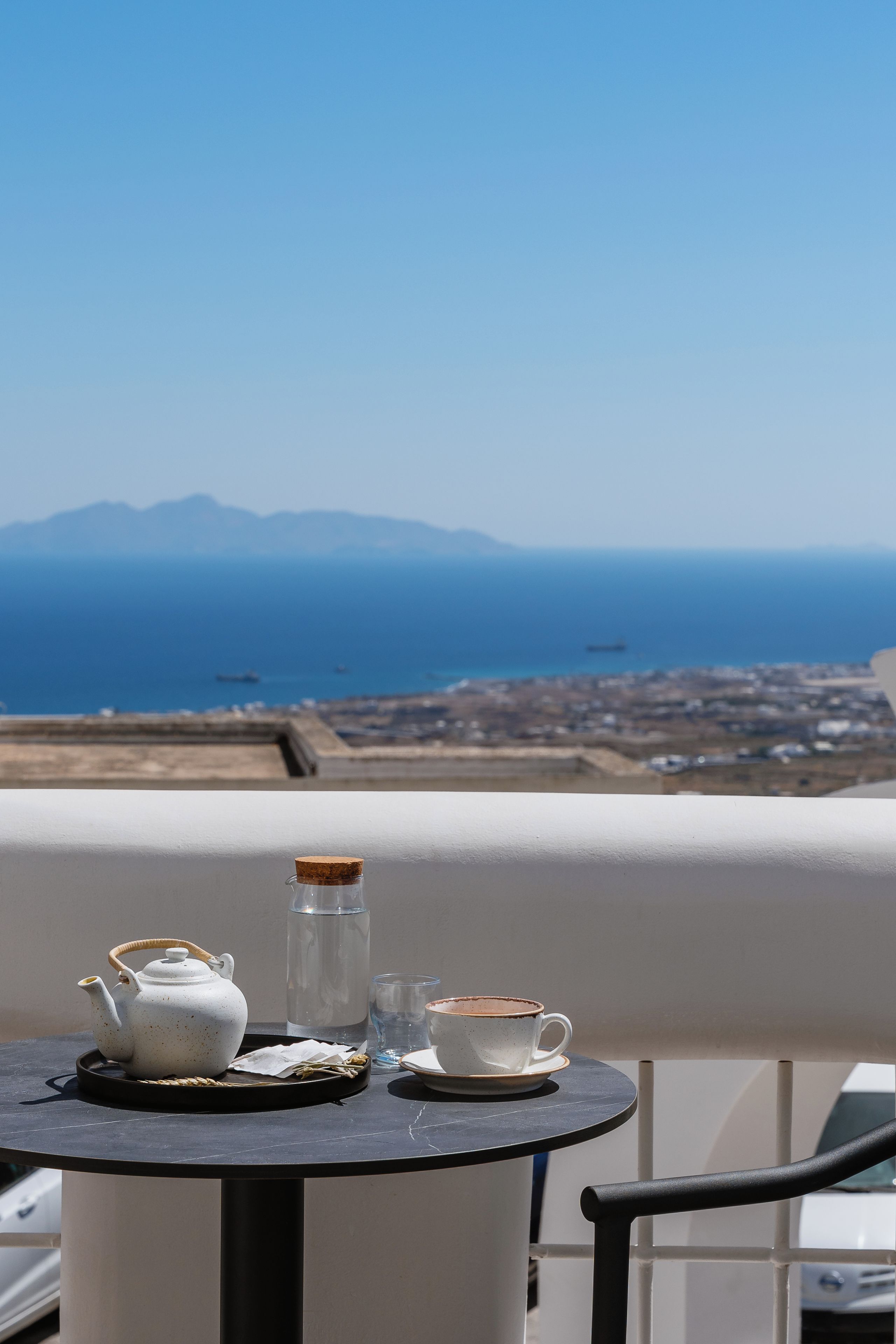 A table with a teapot, a glass, and a cup on a balcony overlooking a coastal view with mountains and a cityscape.