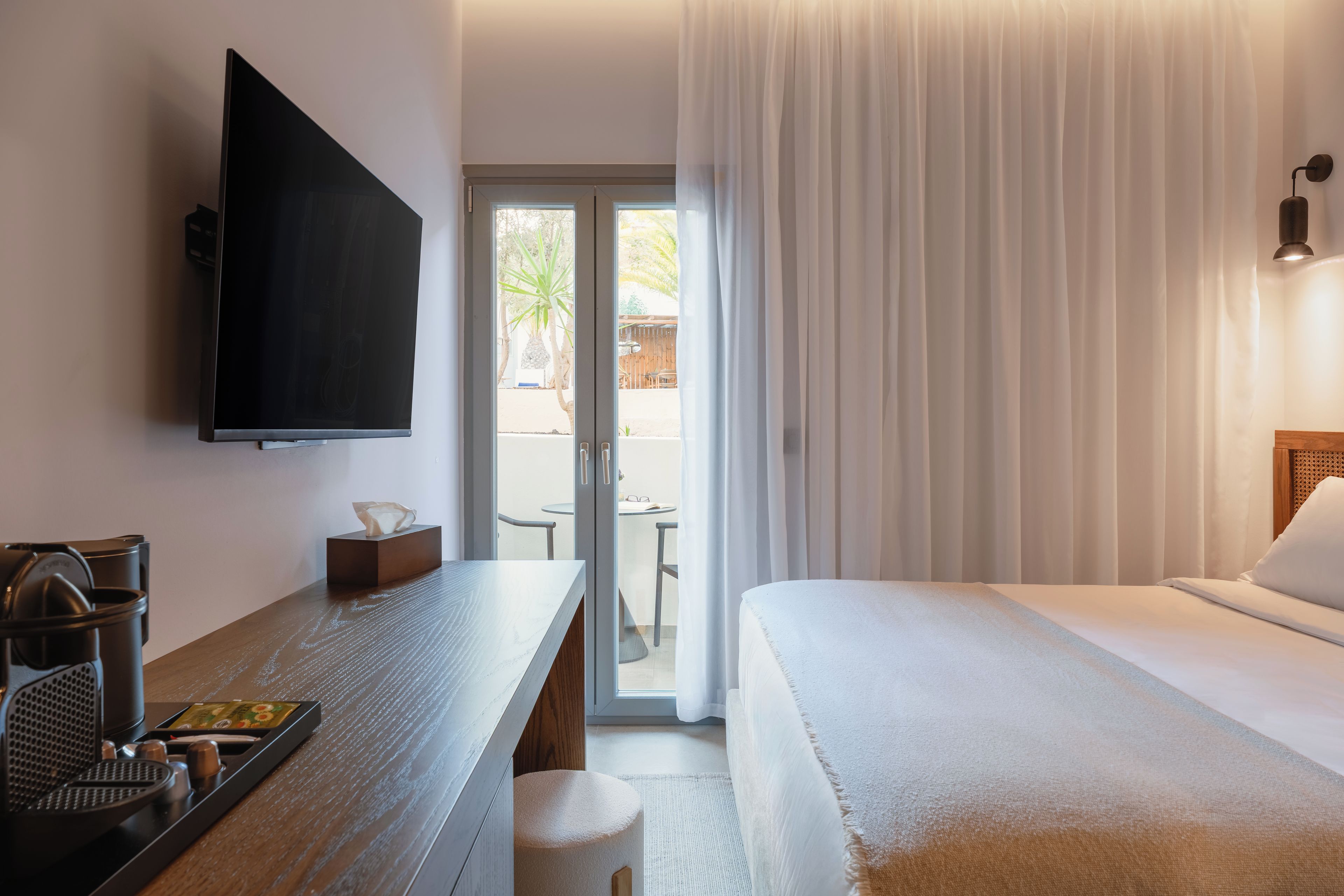 A hotel room with a bed, a desk with a coffee machine, a TV, and a sliding glass door to an outdoor area.