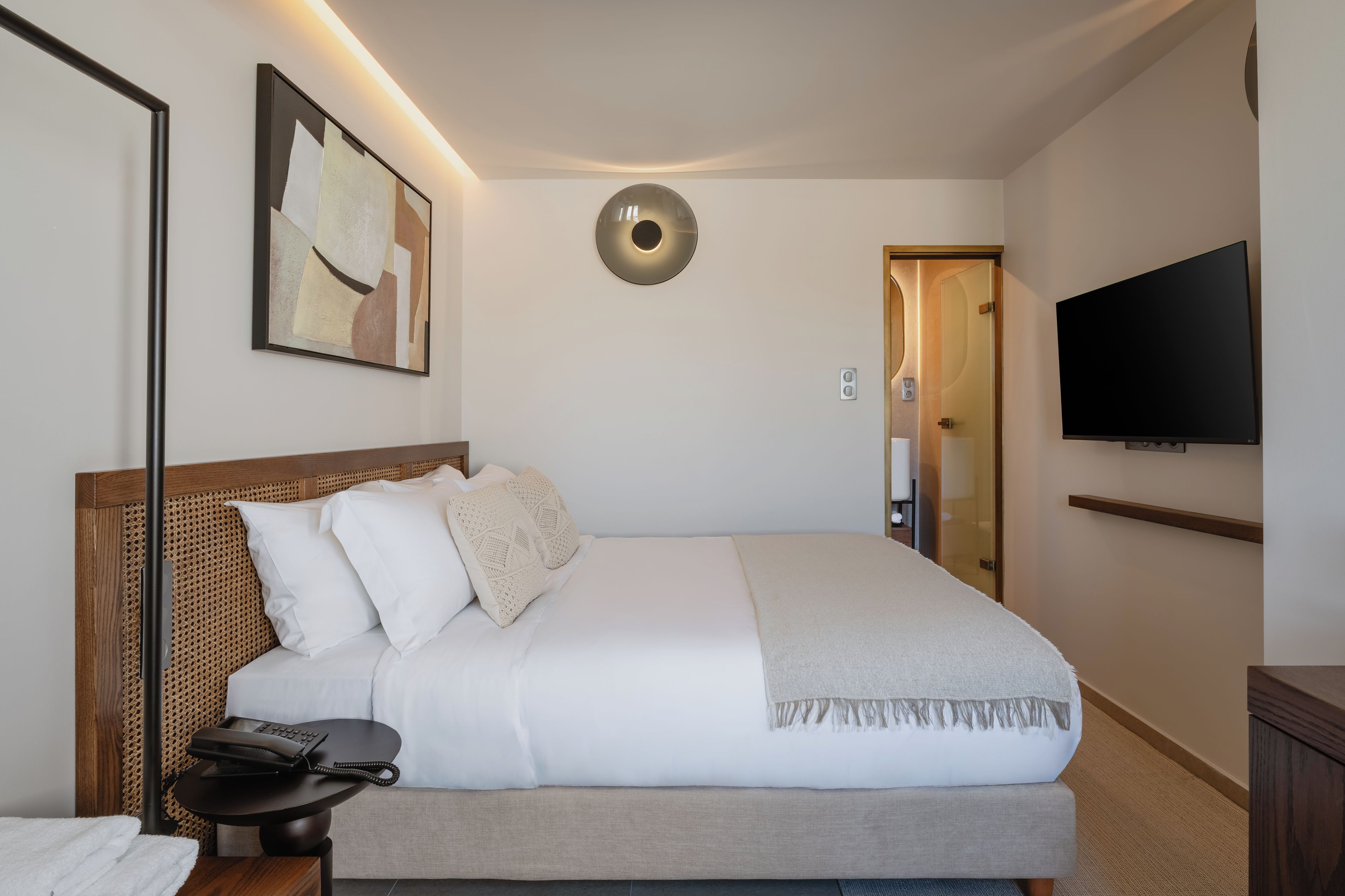 A neatly made bed with a headboard, a bedside table with a telephone, a wall-mounted TV, and a framed abstract painting.