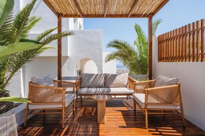 Un espacio al aire libre con mobiliario de mimbre y una mesa de madera, rodeado de paredes blancas y palmeras.
