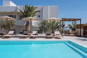Zona de piscina con sillones de descanso y sombrillas, una piscina azul cristalina y un edificio blanco al fondo.
