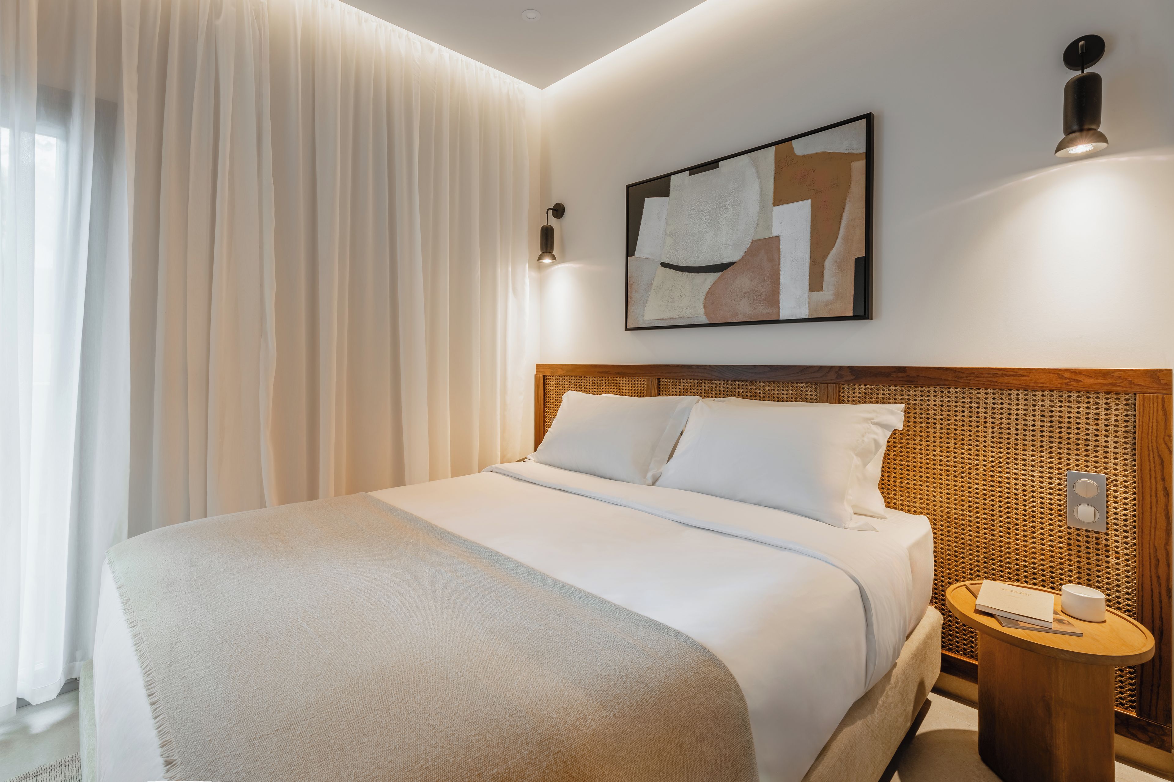 A hotel room with a bed, a nightstand, a wall-mounted light, and a painting.