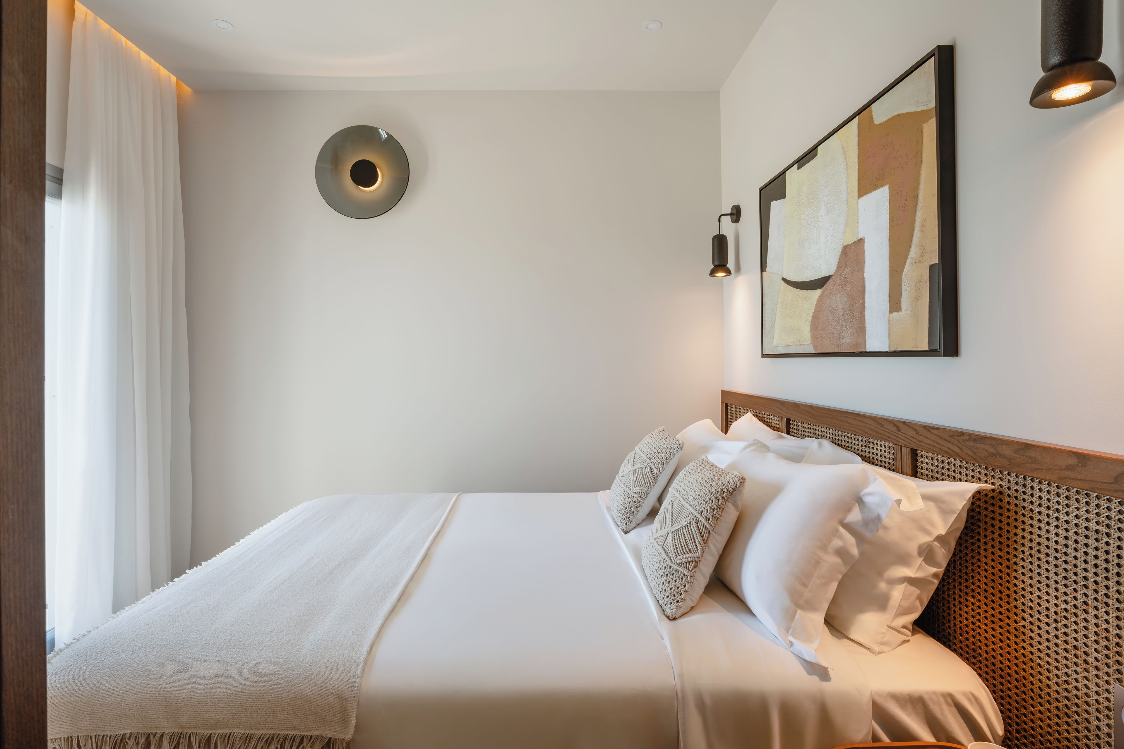 Una cama bien hecha con una colcha blanca y dos almohadones decorativos, una lámpara de pared y una obra de arte abstracta en la pared.