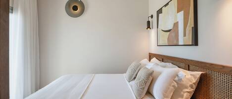 Una cama bien hecha con una colcha blanca y dos almohadones decorativos, una lámpara de pared y una obra de arte abstracta en la pared.