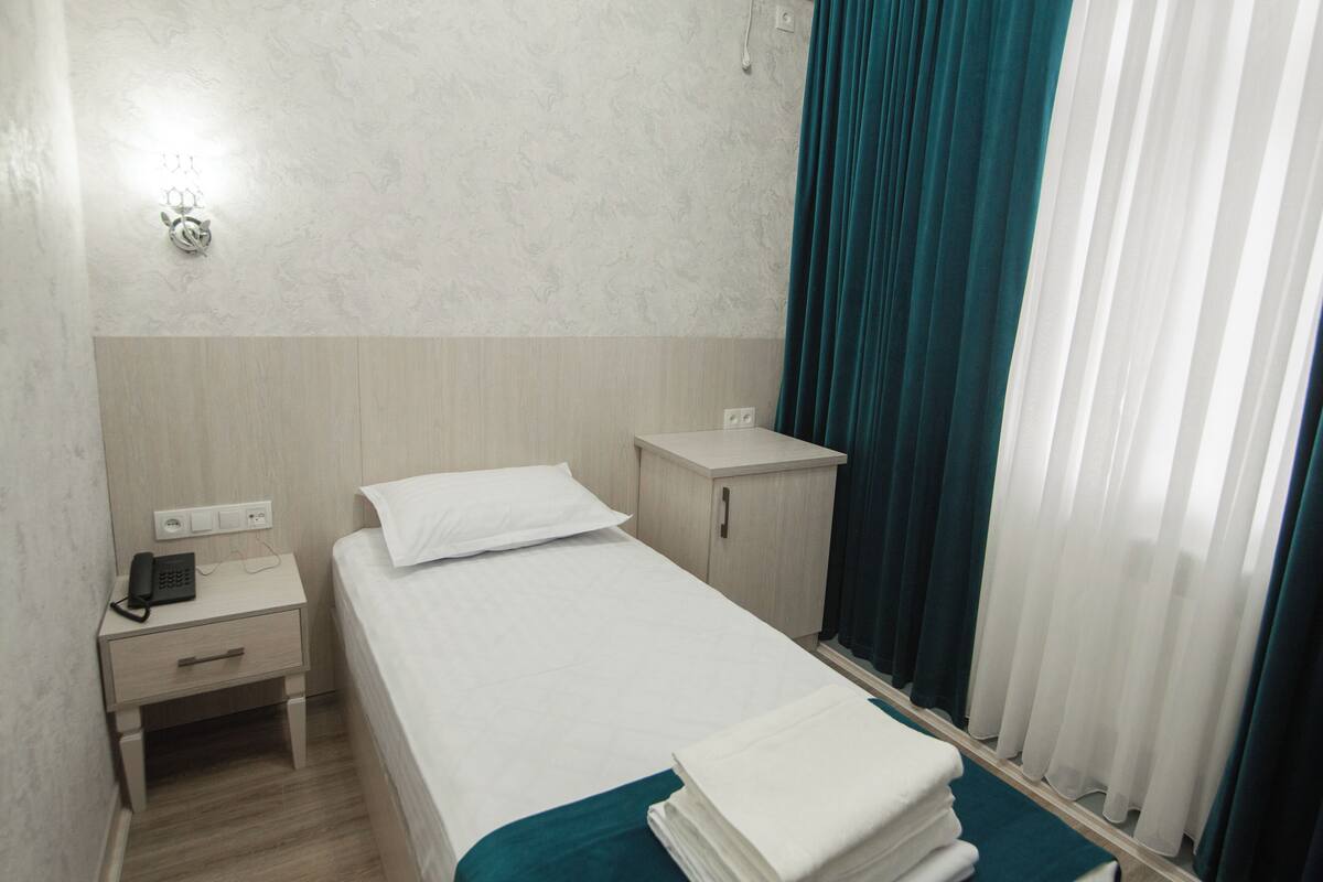 A hotel room with a bed, a nightstand with a phone, a small cabinet, and a window with curtains.