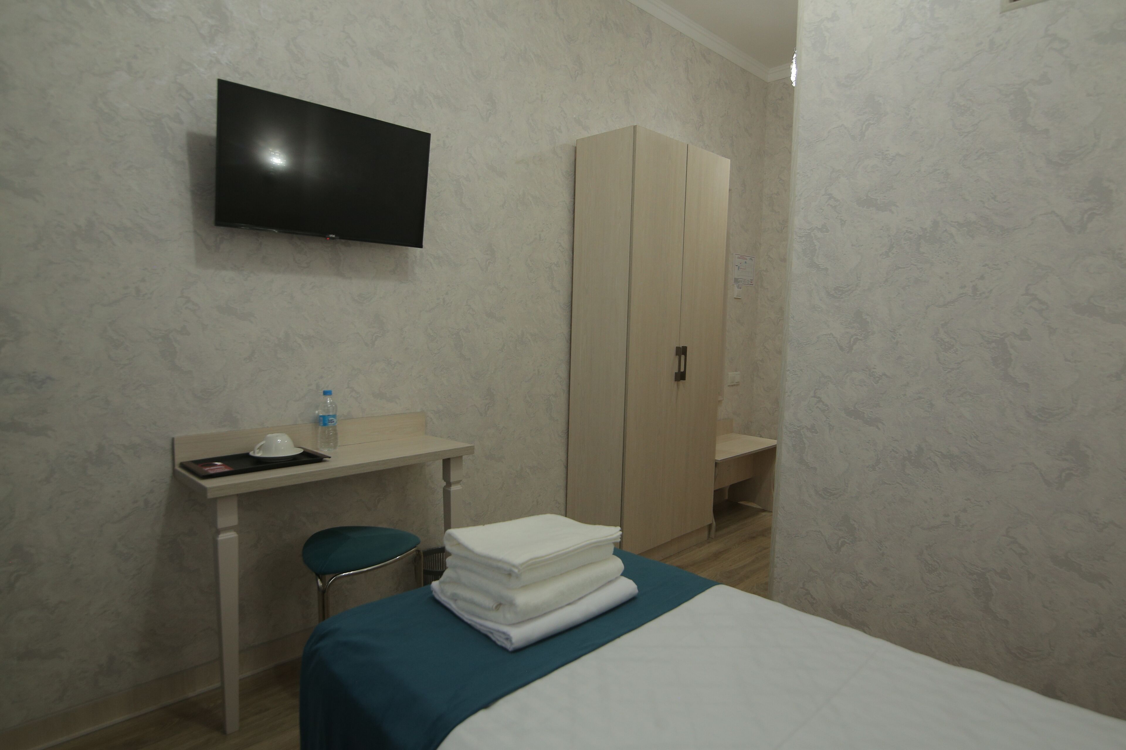 A hotel room with a bed, a TV mounted on the wall, a desk with a chair, and a wardrobe.