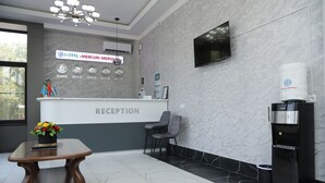 A reception area with a reception desk, chairs, a table with a flower arrangement, a TV mounted on the wall, and a water dispenser. - HOTEL MERCURI MERIDIAN (Bukhara)