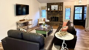 Una sala de estar con un sofá de cuero negro, una mesa de centro, un televisor montado en la pared, una chimenea y un ventilador de techo.