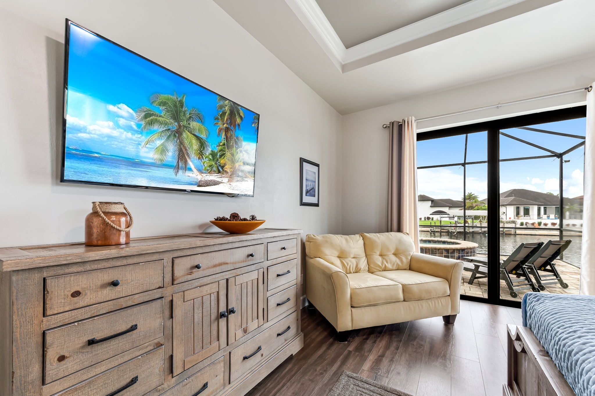 A modern hotel room with a large TV displaying a beach scene, a wooden cabinet with drawers, a beige sofa, and a balcony with a view of the water and buildings.