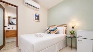 A hotel room with a bed, a bedside table with a lamp, a refrigerator, and a wall-mounted air conditioner.