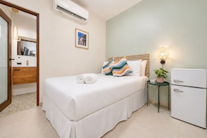A hotel room with a bed, a bedside table with a lamp, a refrigerator, and a wall-mounted air conditioner.