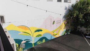 Une fresque colorée orne le mur extérieur d’un bâtiment, un pot de fleurs y est posé, et le ciel est dégagé.