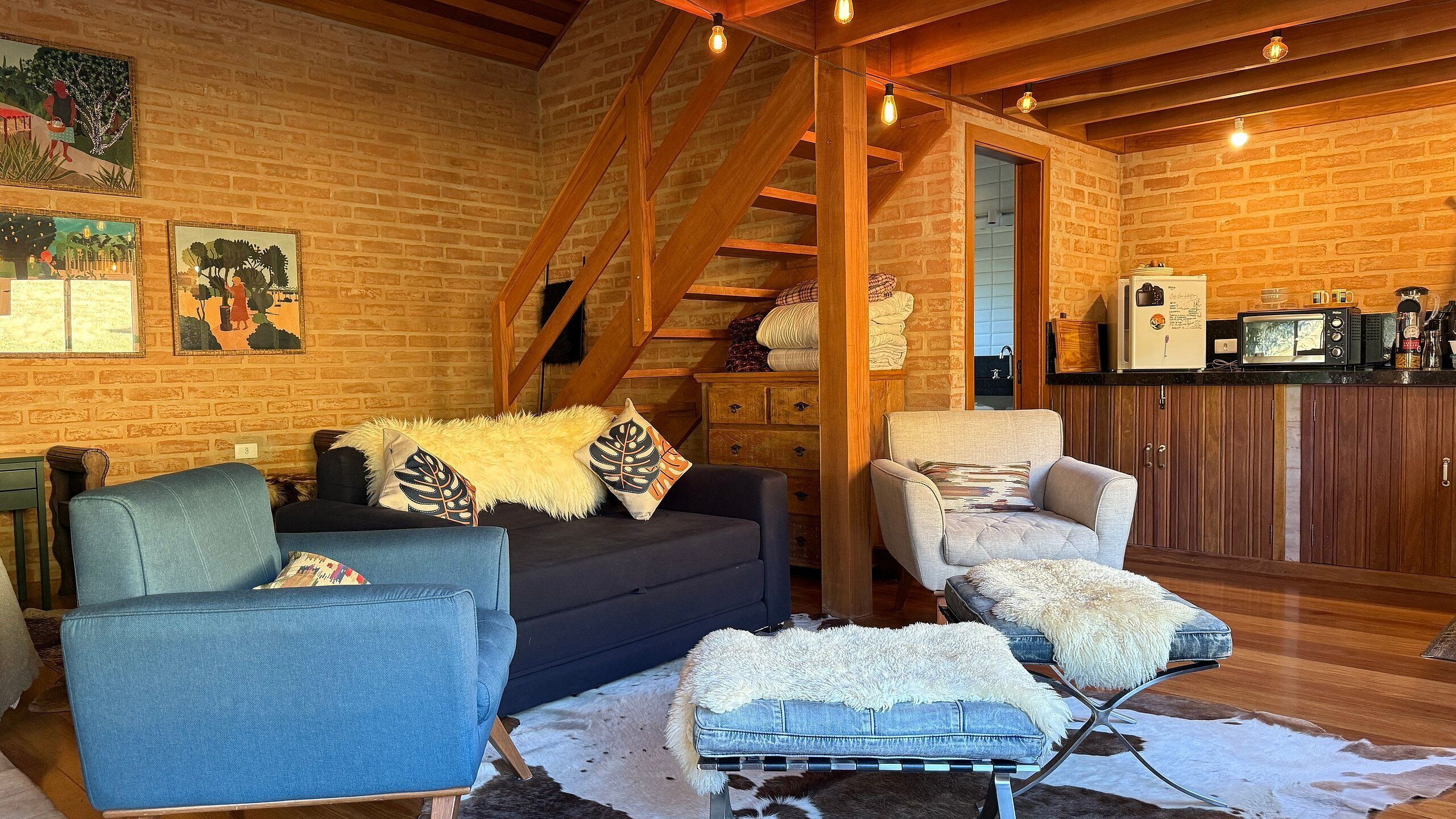 Ein gemütliches Wohnzimmer mit Backsteinwand, einer Treppe, einer Couch, einem Sessel und einer kleinen Küchenzeile.