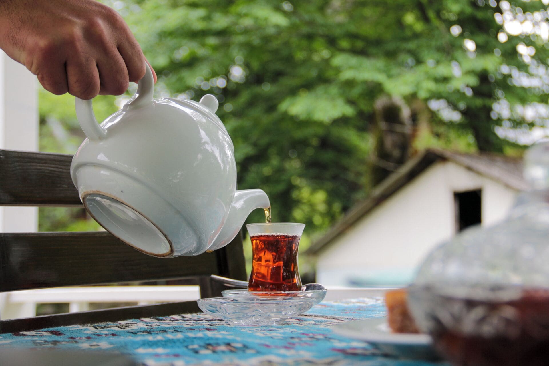 A white teapot pouring tea into a glass cup on a table with a blue patterned tablecloth.