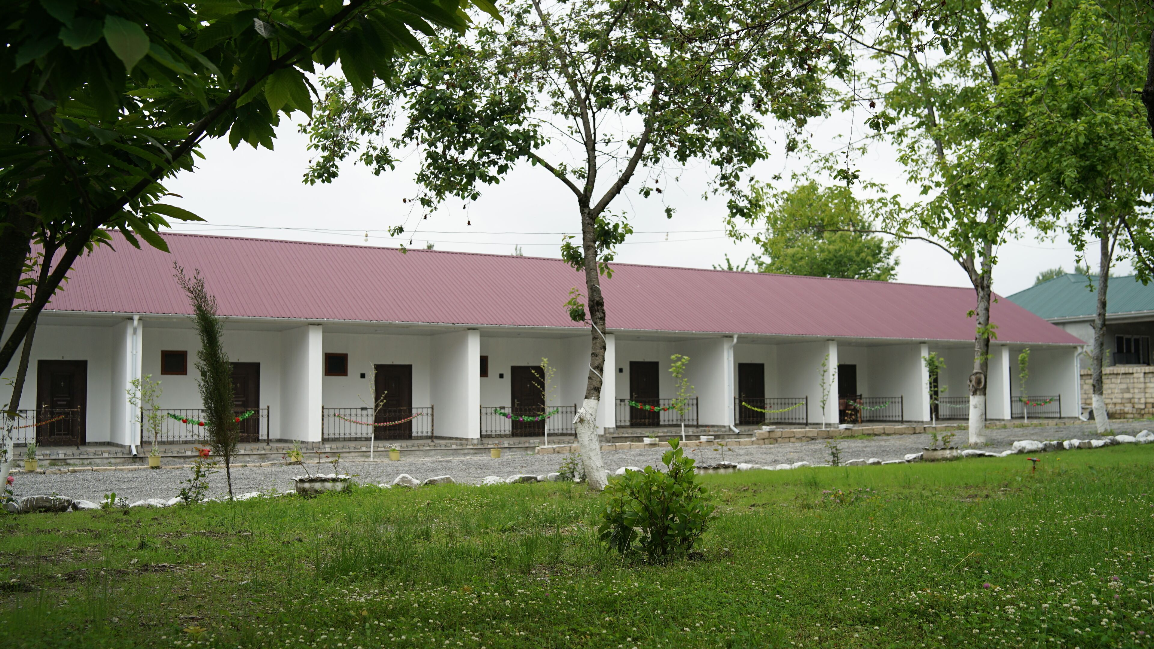 A building with a red roof and white walls, featuring a series of doors and windows, surrounded by a grassy area with trees.