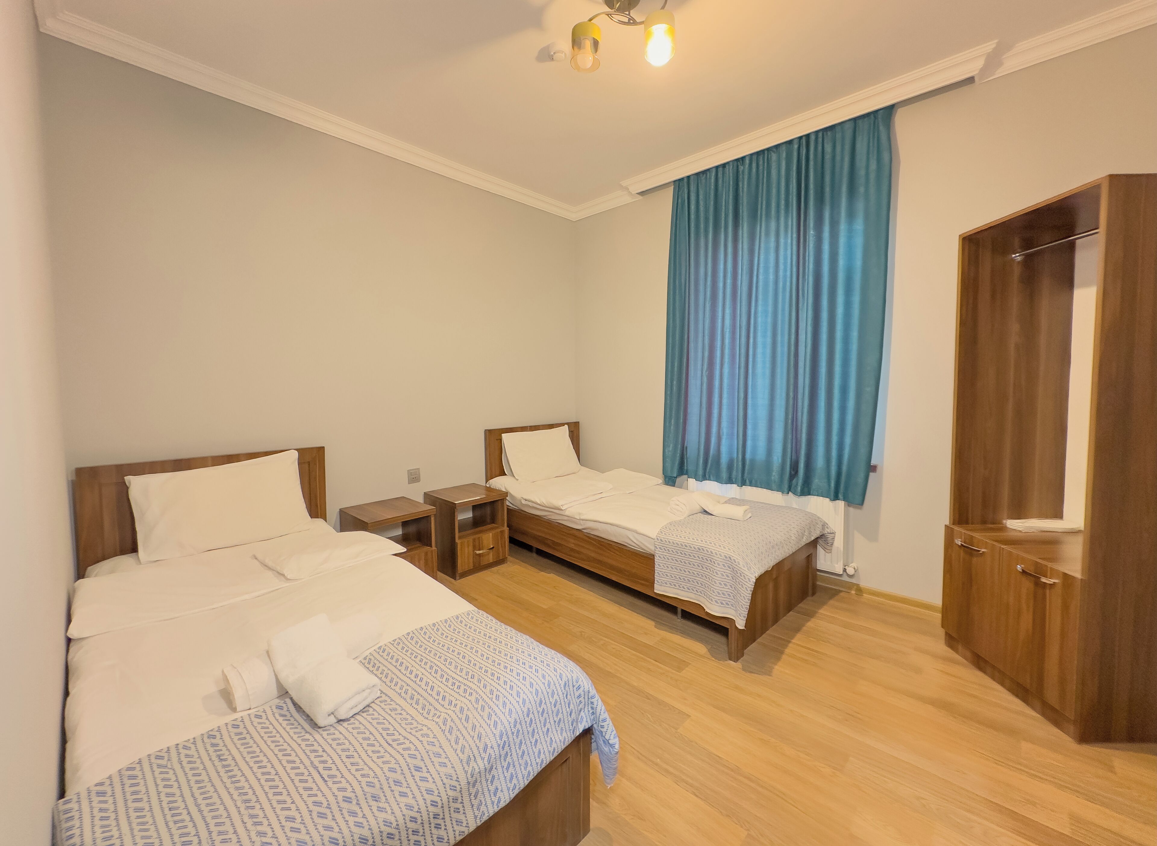 Ein Hotelzimmer mit zwei Einzelbetten, einem Holzschrank und einem großen Fenster mit blauen Vorhängen.