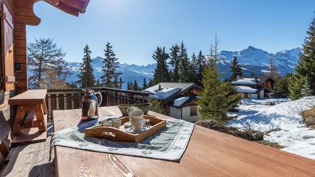 A wooden deck with a table set for tea, overlooking a snowy mountain landscape and traditional buildings.