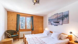 A cozy bedroom with a wooden bed, a chair, a small table, and a window with curtains.