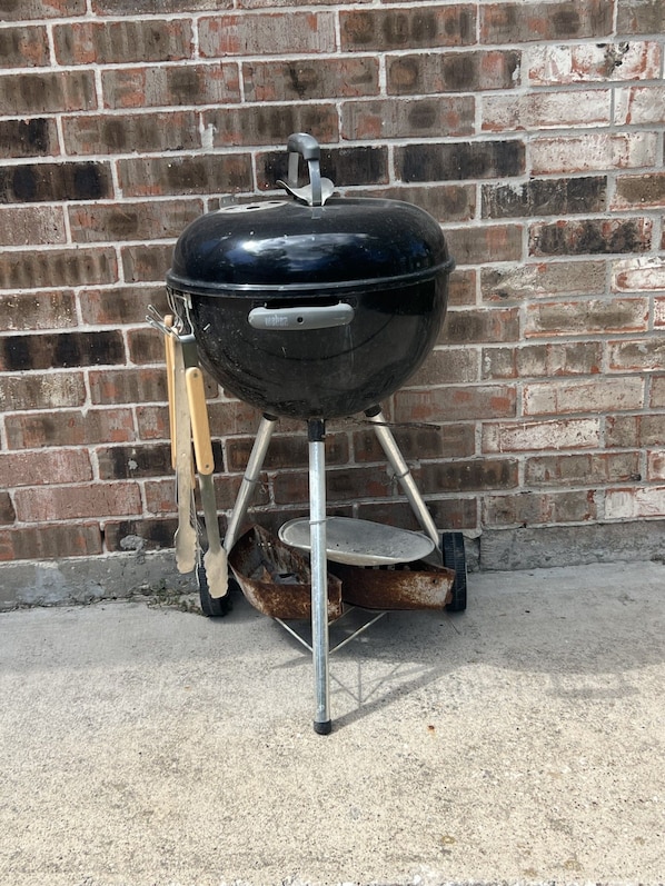 Una parrilla negra para exteriores con soporte metálico y mango de madera, ubicada frente a un muro de ladrillo.