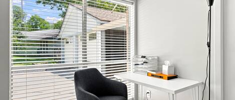 A room with a white desk, a black chair, and a window with blinds.