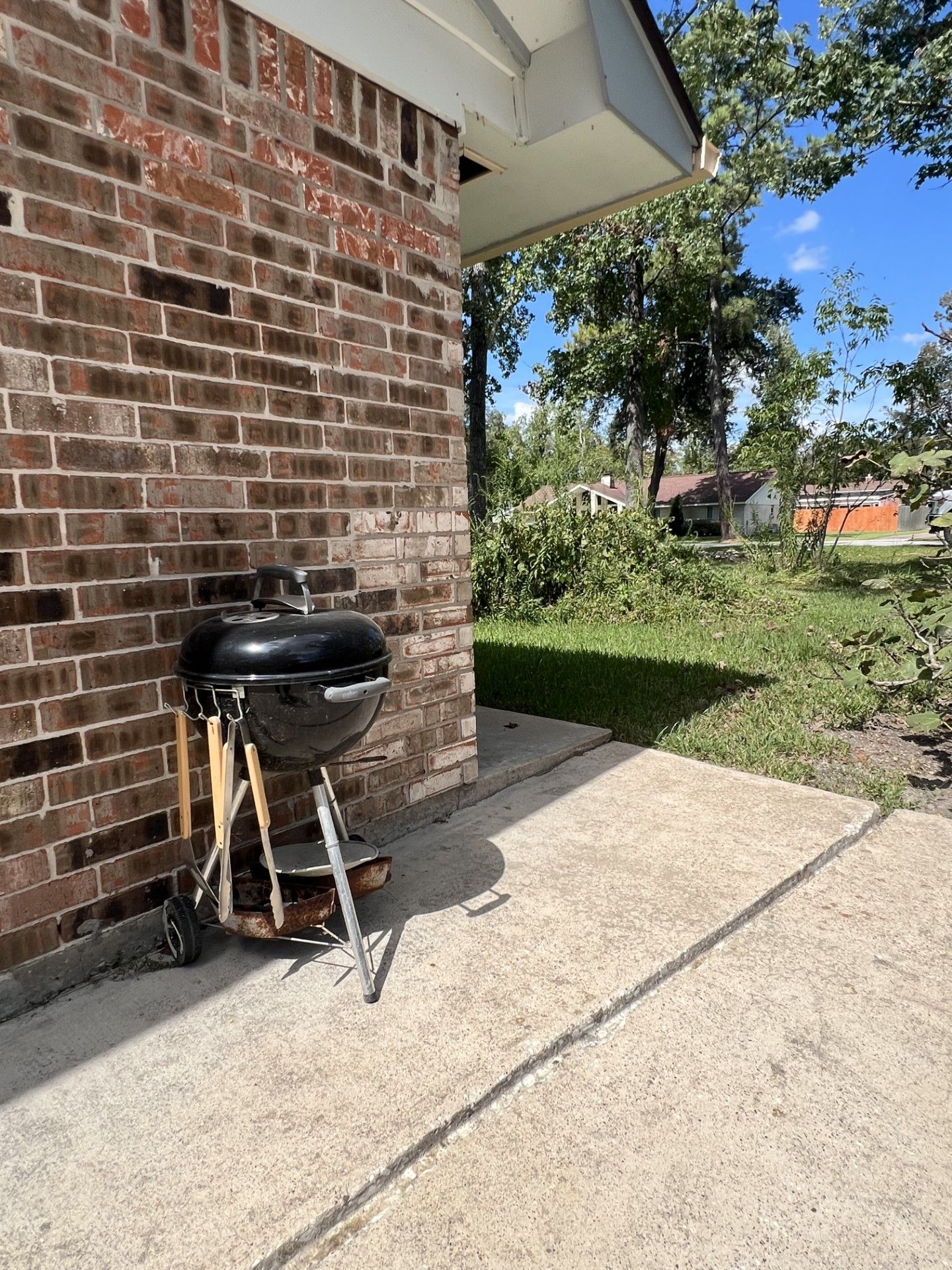 A barbecue grill positioned against a brick wall, with a clear view of the surrounding yard and a house in the background.