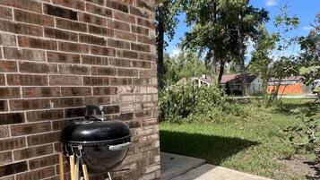 A barbecue grill positioned against a brick wall, with a clear view of the surrounding yard and a house in the background.