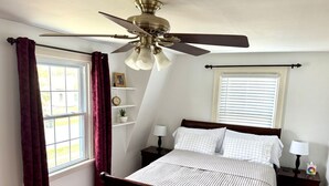 A bedroom with a bed, a ceiling fan, a window with curtains, a bedside table with a lamp, and a nightstand with a lamp.