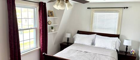 A bedroom with a bed, a ceiling fan, a window with curtains, a bedside table with a lamp, and a nightstand with a lamp.