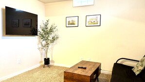A living room with a flat-screen TV, two framed pictures, a small wooden table, a potted plant, and a black sofa.