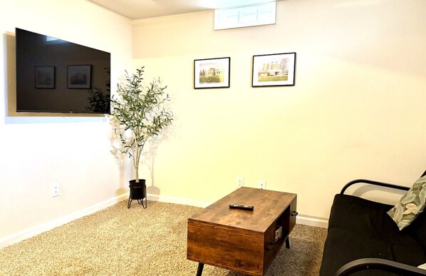 Un salon avec un téléviseur à écran plat, deux tableaux encadrés, une petite table en bois, une plante en pot et un canapé noir.
