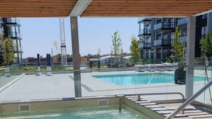A rooftop swimming pool with clear water, surrounded by modern buildings and a covered area with wooden beams.