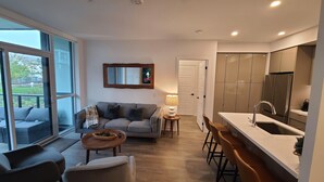 A modern living room with a sofa, a coffee table, and a dining area with bar stools. There is a kitchen with a sink and a refrigerator.