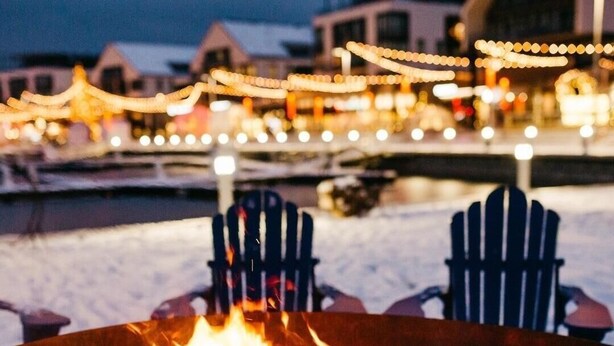 A fire pit with burning logs, surrounded by Adirondack chairs, and a snowy landscape in the background.