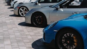 A row of parked sports cars, including a blue Porsche GTS, on a paved surface with modern buildings in the background.