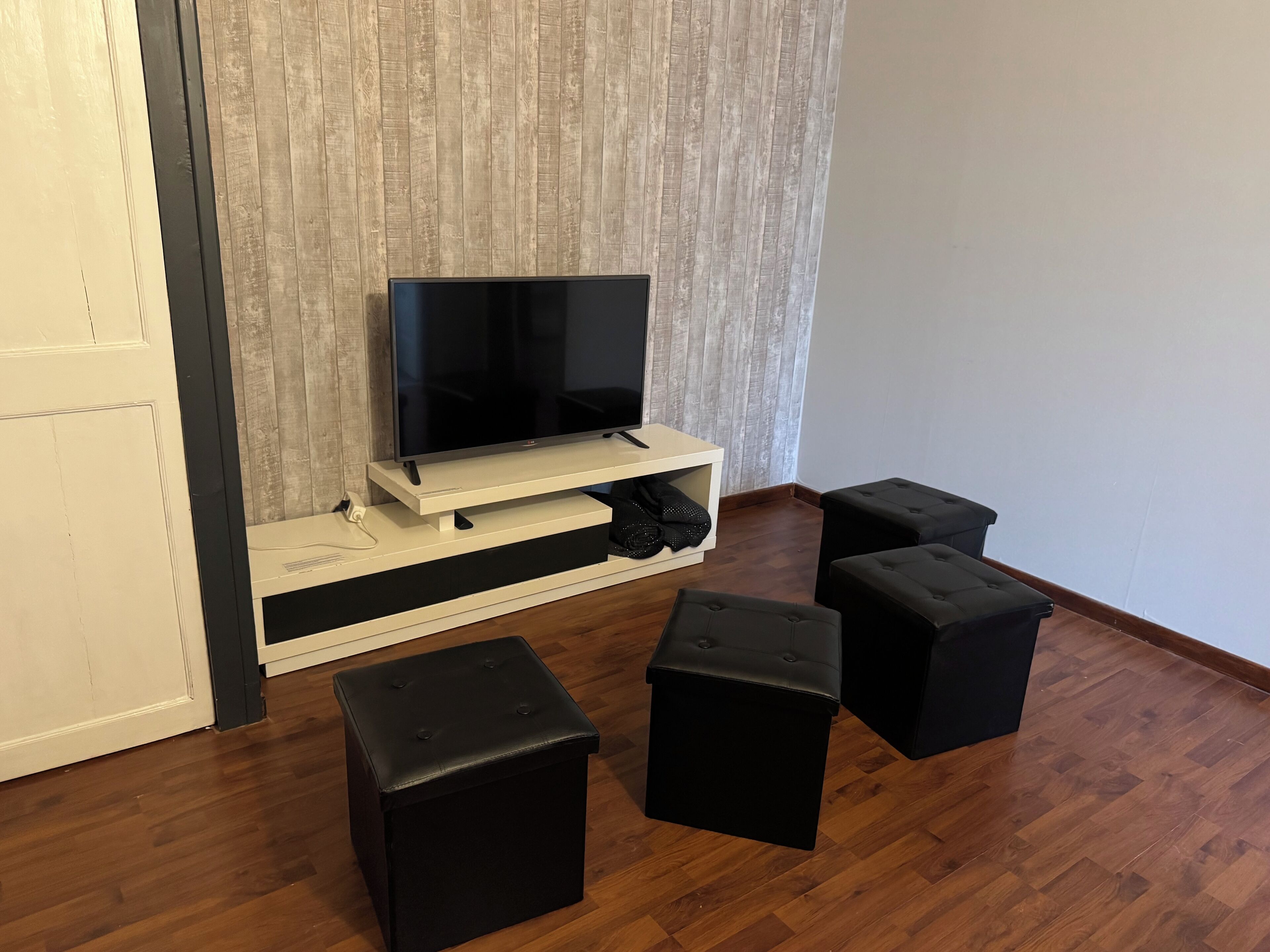 Un soggiorno con televisore su un supporto in legno, poggiapiedi neri e una porta bianca.