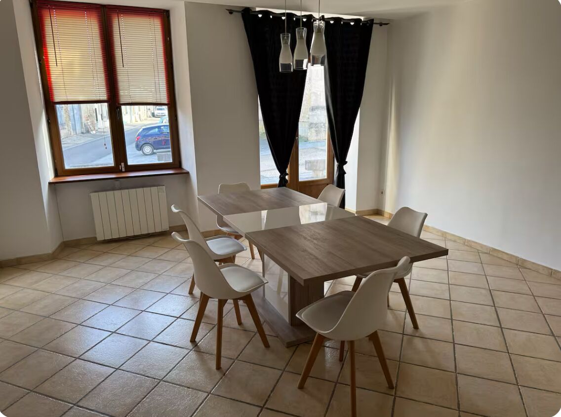 Een eetgedeelte met een houten tafel en witte stoelen, een radiator onder het raam en uitzicht op de straat.