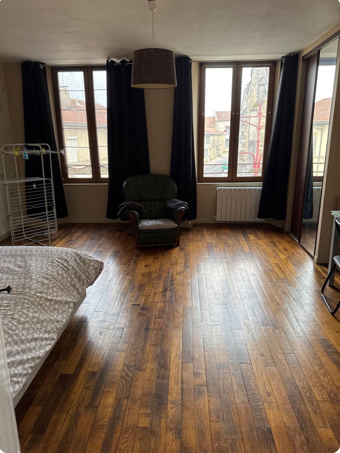 A room with wooden flooring, a single chair, a bed, and large windows with curtains.