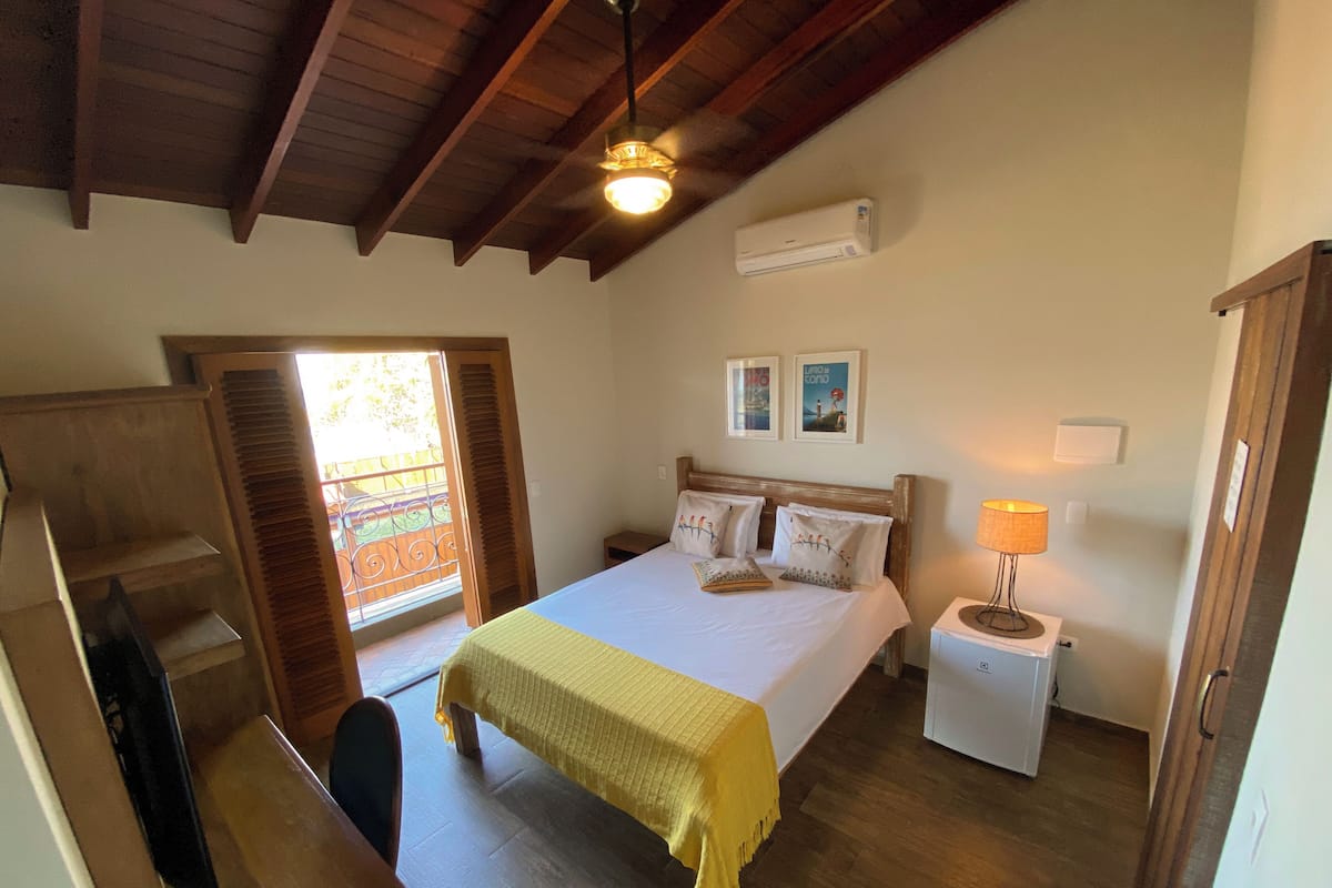 Un dormitorio con cama, mesita de noche, lámpara, ventilador de techo y ventana con contraventanas.