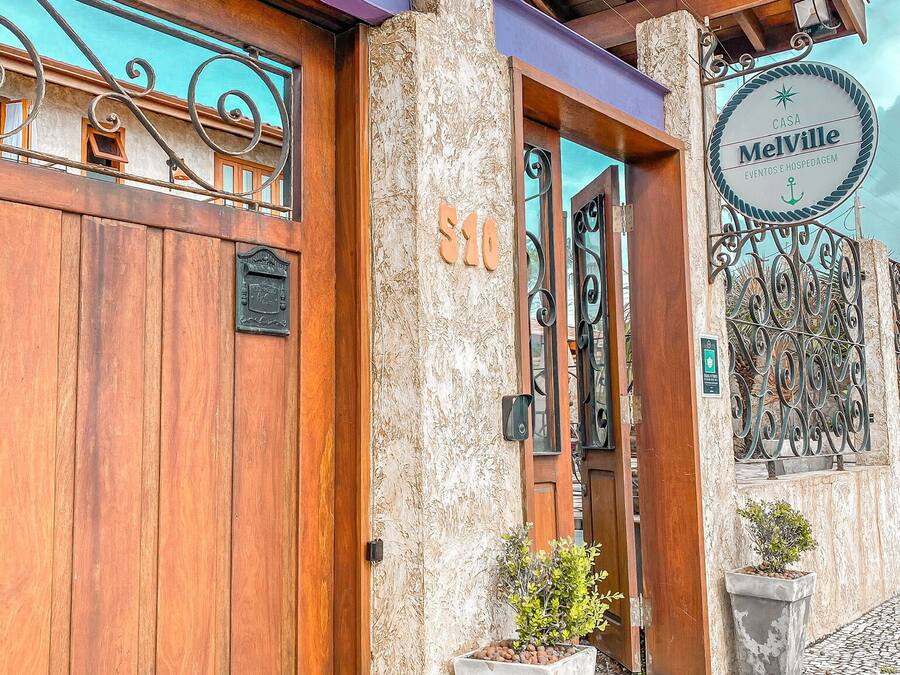 Entrada de un edificio con puerta de madera, un pilar de piedra y un cartel que dice "Casa MelVille Eventos e Hospedagem".