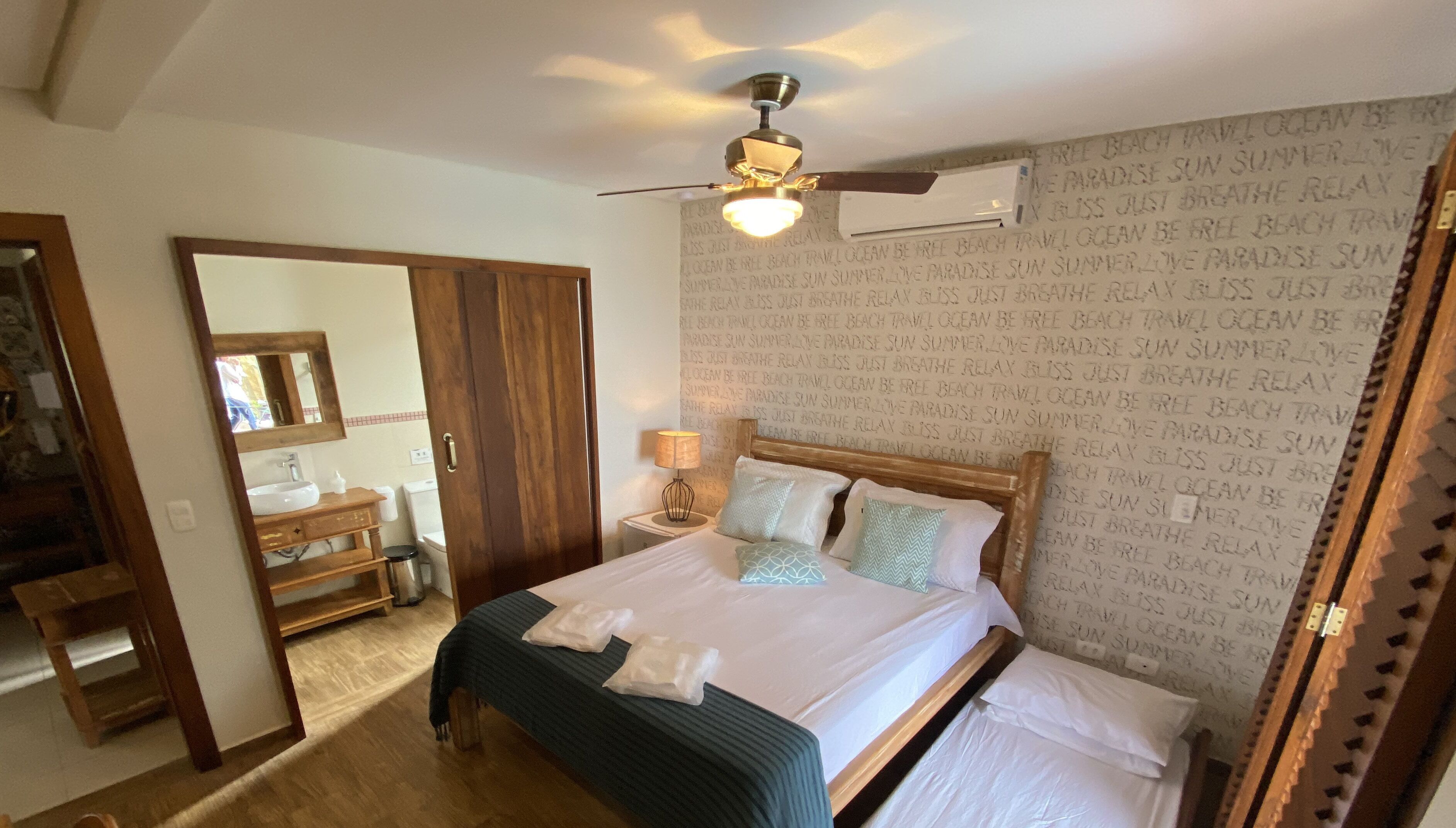 A bedroom with a bed, a ceiling fan, a nightstand, a mirror, and a bathroom visible through an open door.