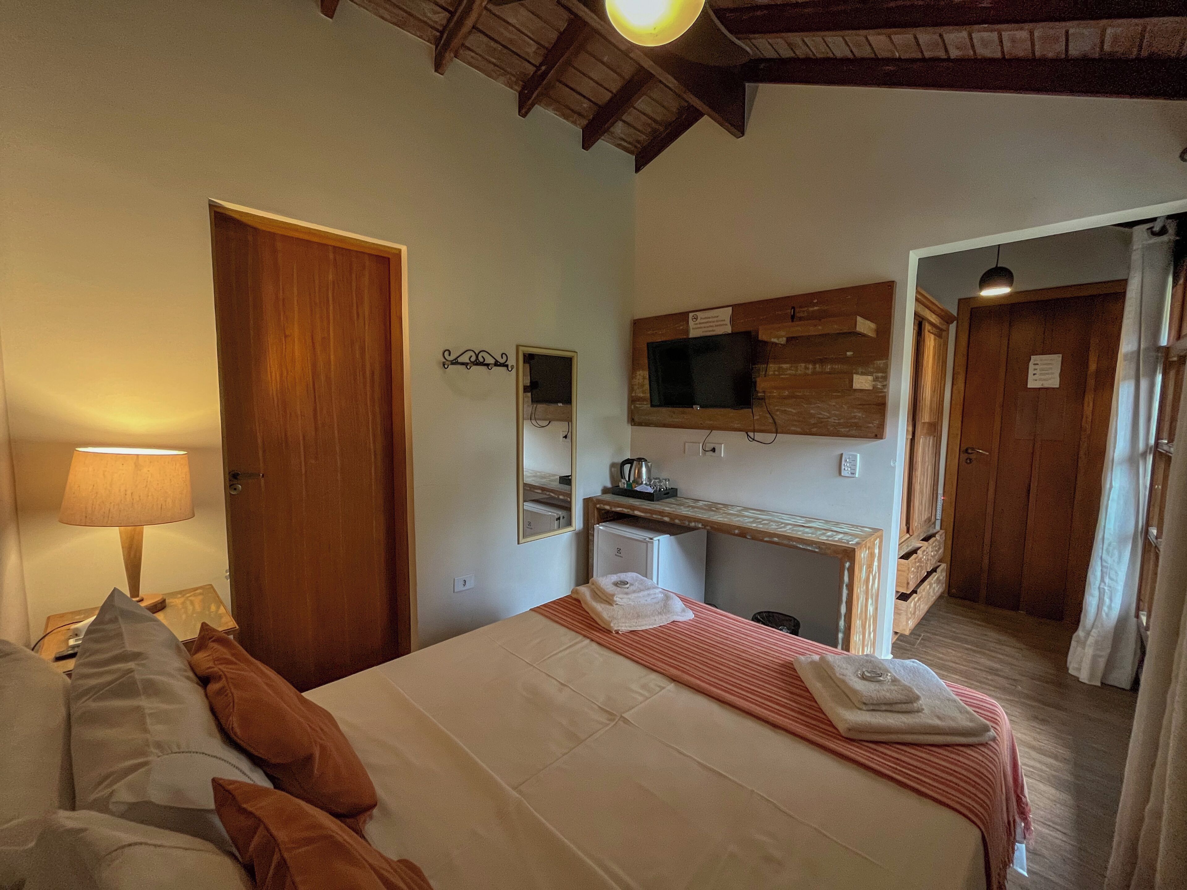 A hotel room with a bed, a desk, a TV mounted on the wall, and a wooden ceiling.