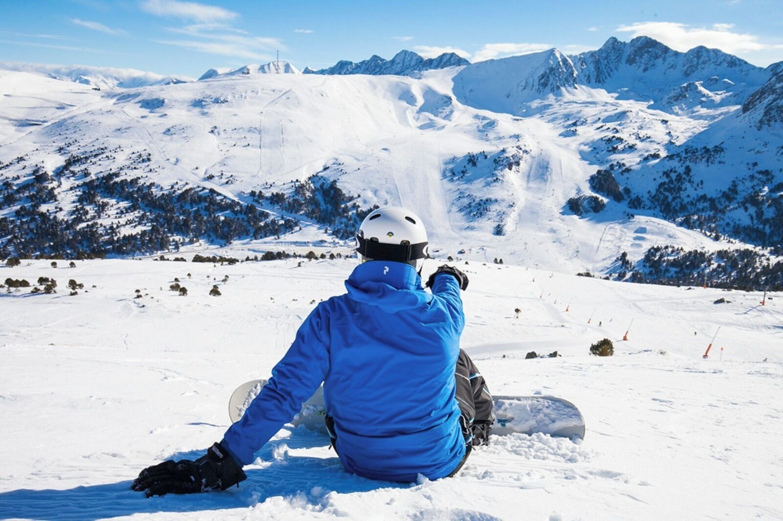 A person sitting on a snow-covered slope with a snowboard, overlooking a snowy mountain range.