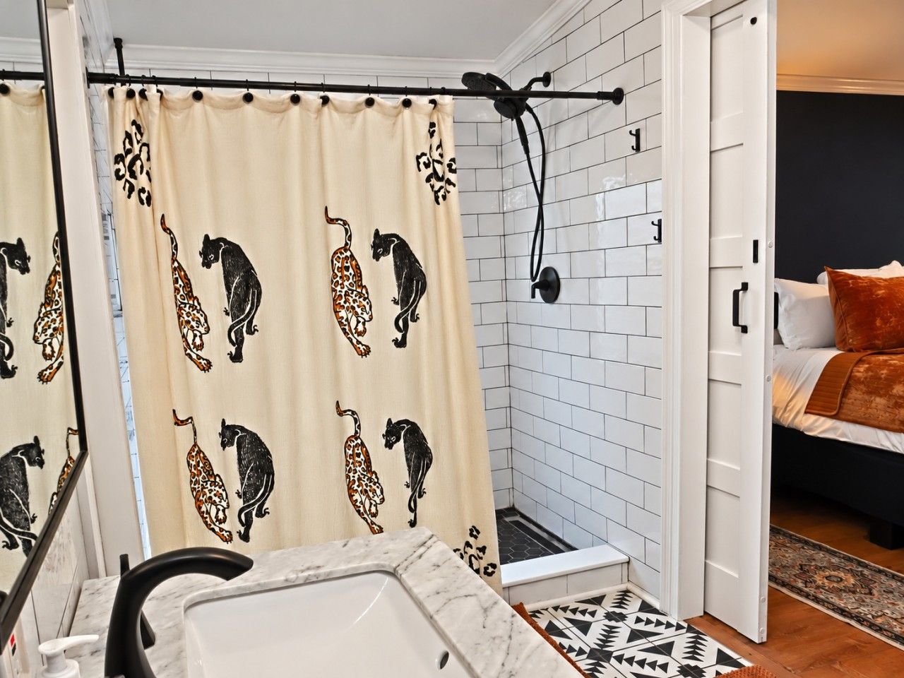 Un baño con cortina de ducha estampada con diseños de animales, pared de azulejos blancos, un cabezal de ducha y una cama visible con un tapete estampado en la habitación contigua.